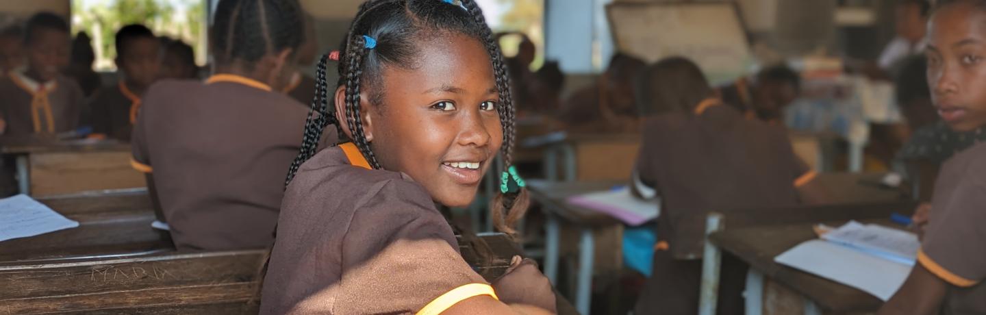 A young girl sitting at a desk in a classroom, with a big smile on her face