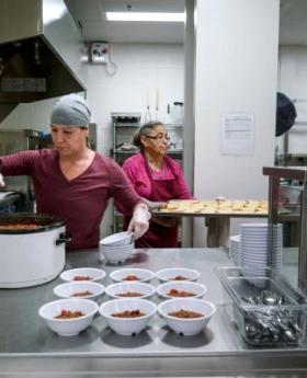 Two women in a commercial kitchen serve food into bowls.