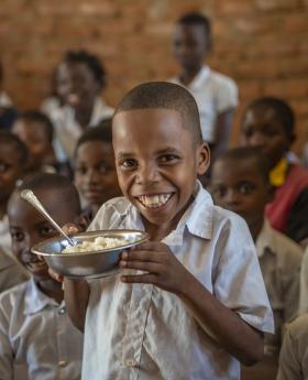 Mignon holds a bowl of food and smiles at the camera, with his friends in the background